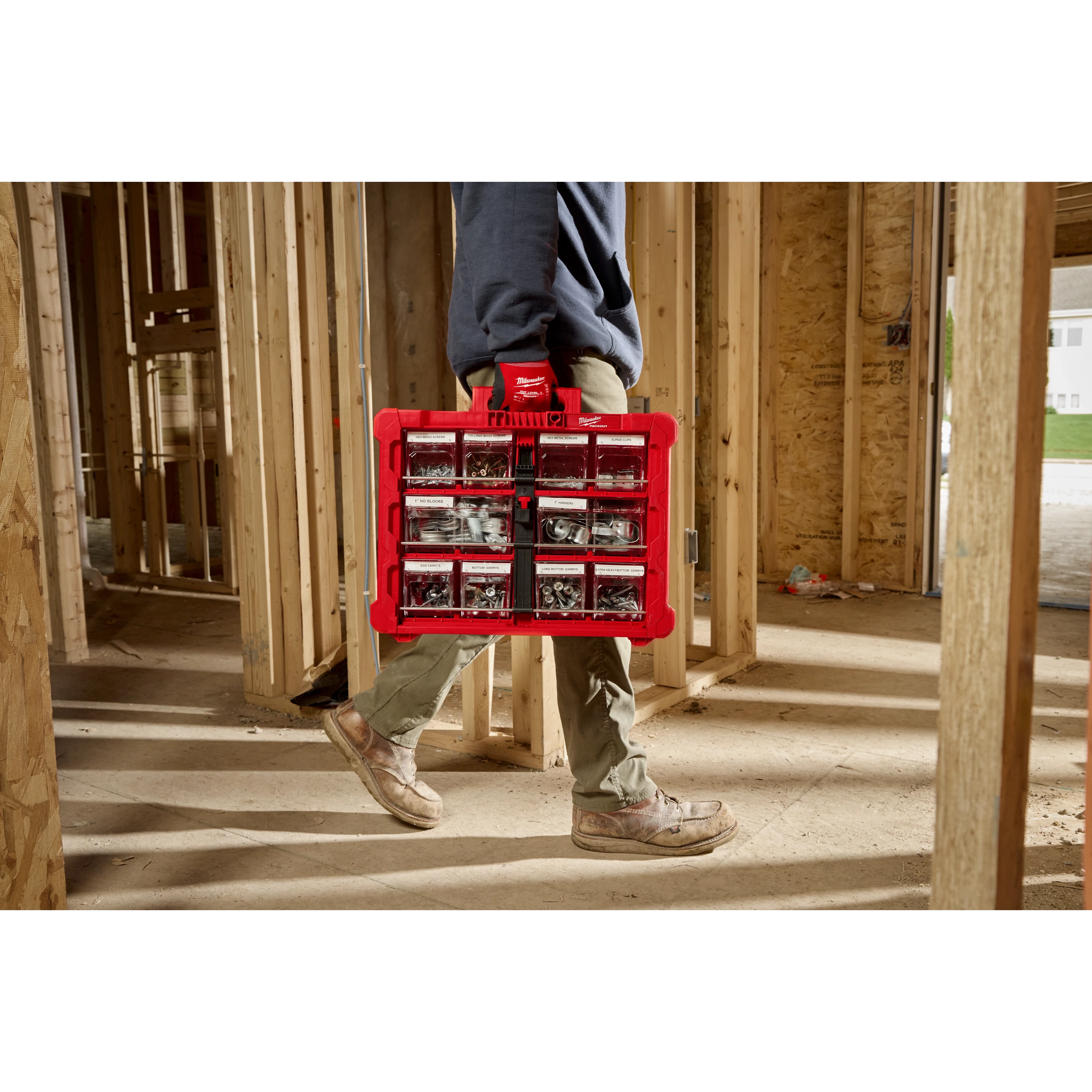 A person in work boots and green pants carries a red plastic organizer box filled with various small tools and hardware. The individual is walking through a wooden framed construction site, with partially completed walls and beams visible. Bright sunlight streams through the space.