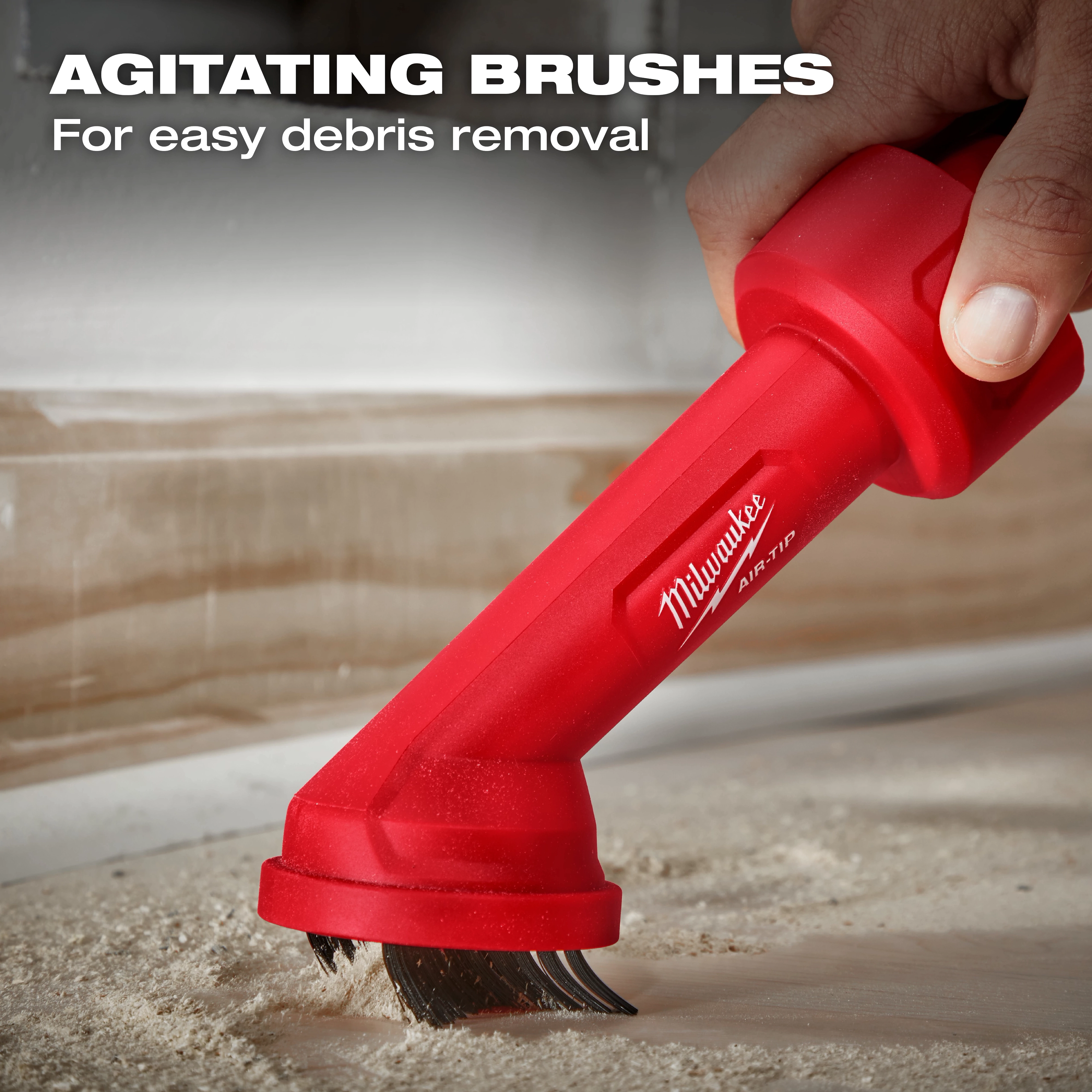 A person uses the AIR-TIP™ Cross Brush Tool by Milwaukee Tool. The tool features agitating brushes designed for easy debris removal, as shown by the sweeping motion along a dusty surface near the edge of a wall. Text reads "AGITATING BRUSHES For easy debris removal".