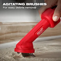 A person uses the AIR-TIP™ Cross Brush Tool by Milwaukee Tool. The tool features agitating brushes designed for easy debris removal, as shown by the sweeping motion along a dusty surface near the edge of a wall. Text reads "AGITATING BRUSHES For easy debris removal".