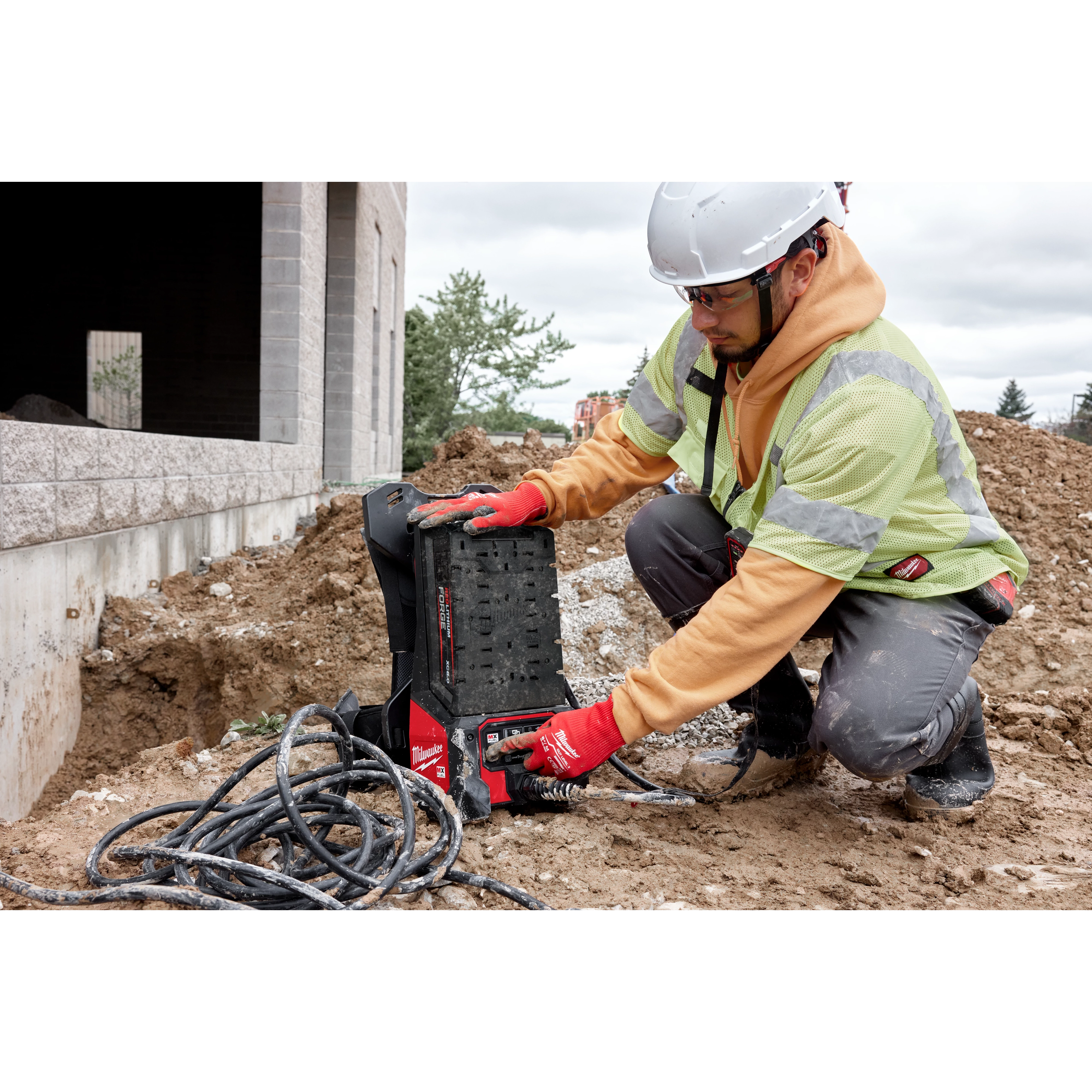 A person in construction gear uses the MX FUEL™ Portable Pump Power Base and MX FUEL™ 1HP 2" Submersible Pump Kit at a construction site. The equipment is placed on the ground with cables lying around it. The environment includes dirt and a partially constructed building.