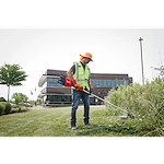 A person in a safety vest and gloves trims grass with a weed cutter outside a modern building.