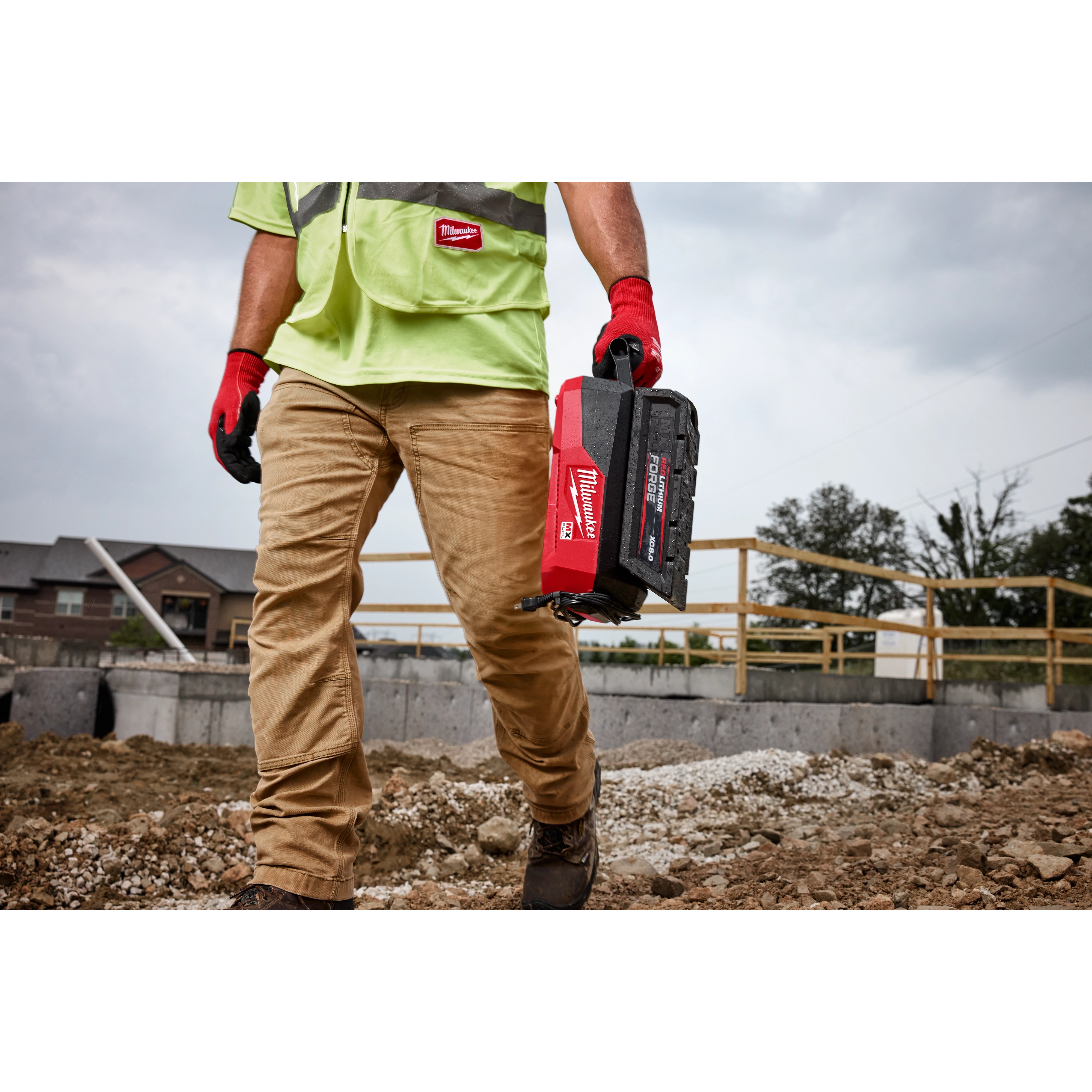 Construction worker carrying a Milwaukee toolbox, wearing safety gear, on a rocky construction site with buildings in the background.