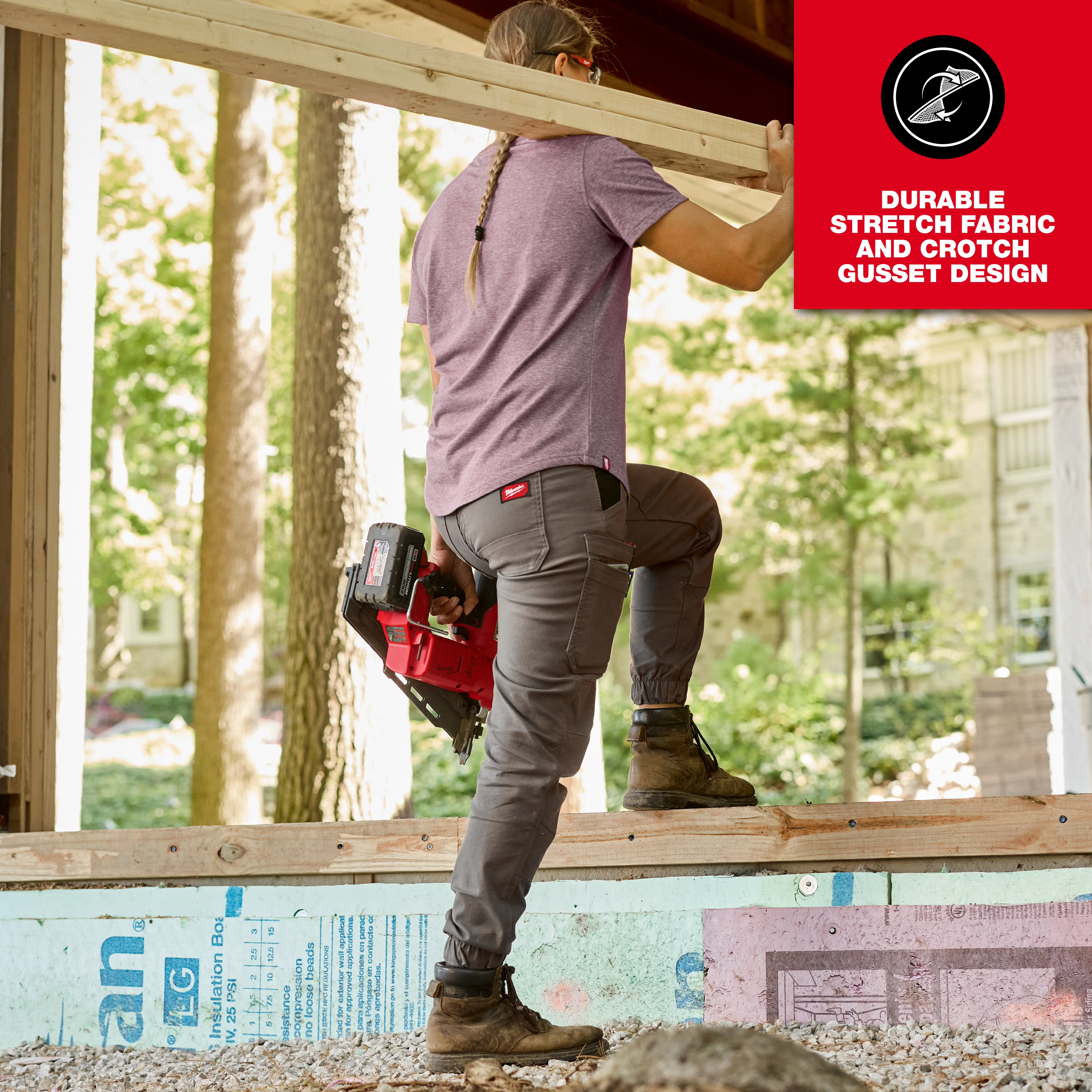 A woman wearing gray Women's GRIDIRON™ Double Knee Jogger Pants and a purple shirt ascends a wooden structure while carrying a power tool. The image highlights the pants' durable stretch fabric and crotch gusset design. A red box with text in the top right corner emphasizes these features.