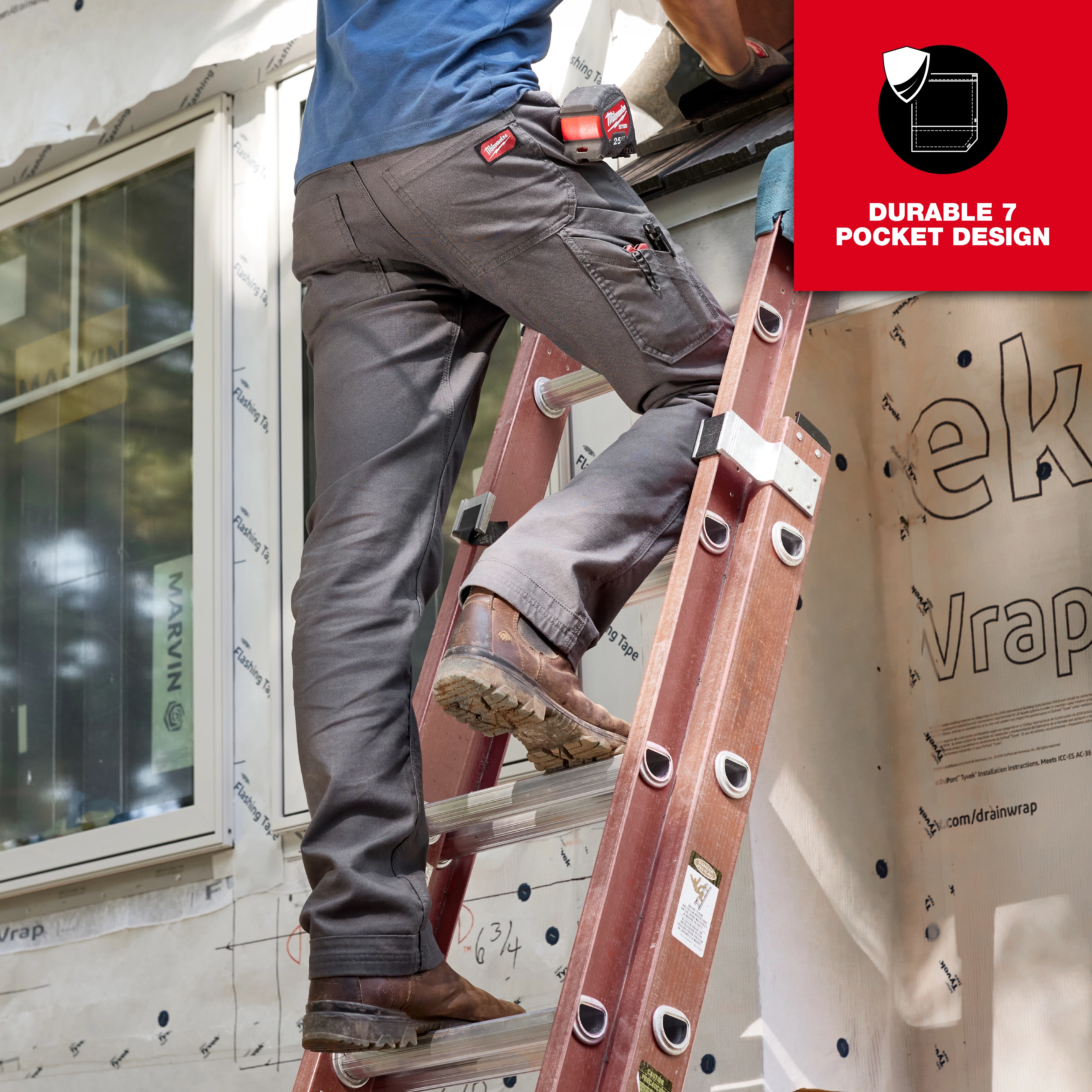 A man wearing gray Work Pants with a durable 7-pocket design is climbing a red ladder outside a building under construction. The pants have multiple pockets and tools attached, highlighting their practicality for work environments.