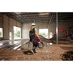 A person wearing a hard hat and protective gear carrying an MX FUEL™ Portable Pump Power Base and an MX FUEL™ 1HP 2" Submersible Pump Kit across a muddy construction site. The scene includes the interior of an unfinished building and construction equipment in the background.