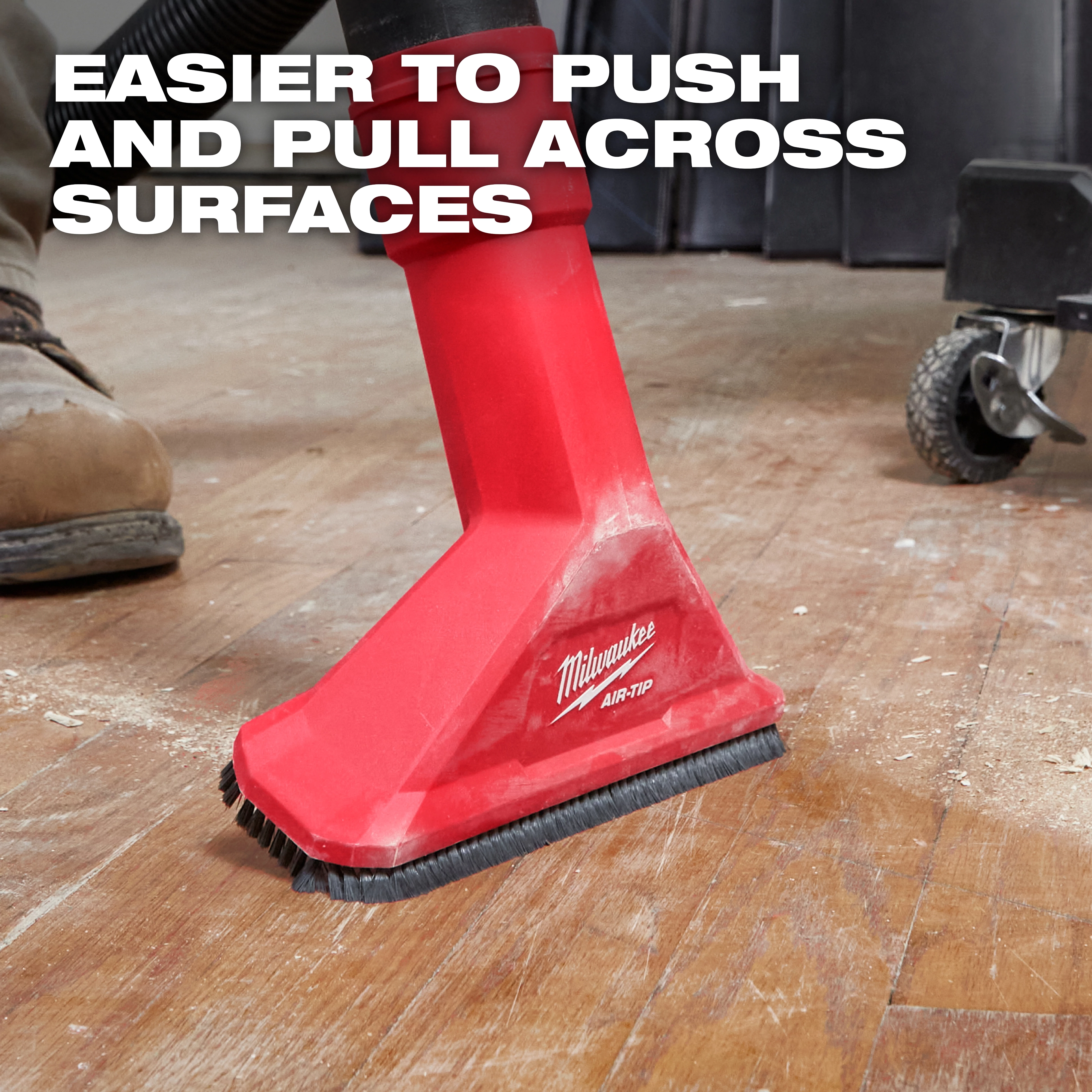 The image shows a red Milwaukee AIR-TIP™ 2-1/2" Rocking Utility Nozzle with Brushes being used on a wooden floor. The nozzle is attached to a vacuum and has brushes at its base. Text on the image reads, "Easier to push and pull across surfaces."