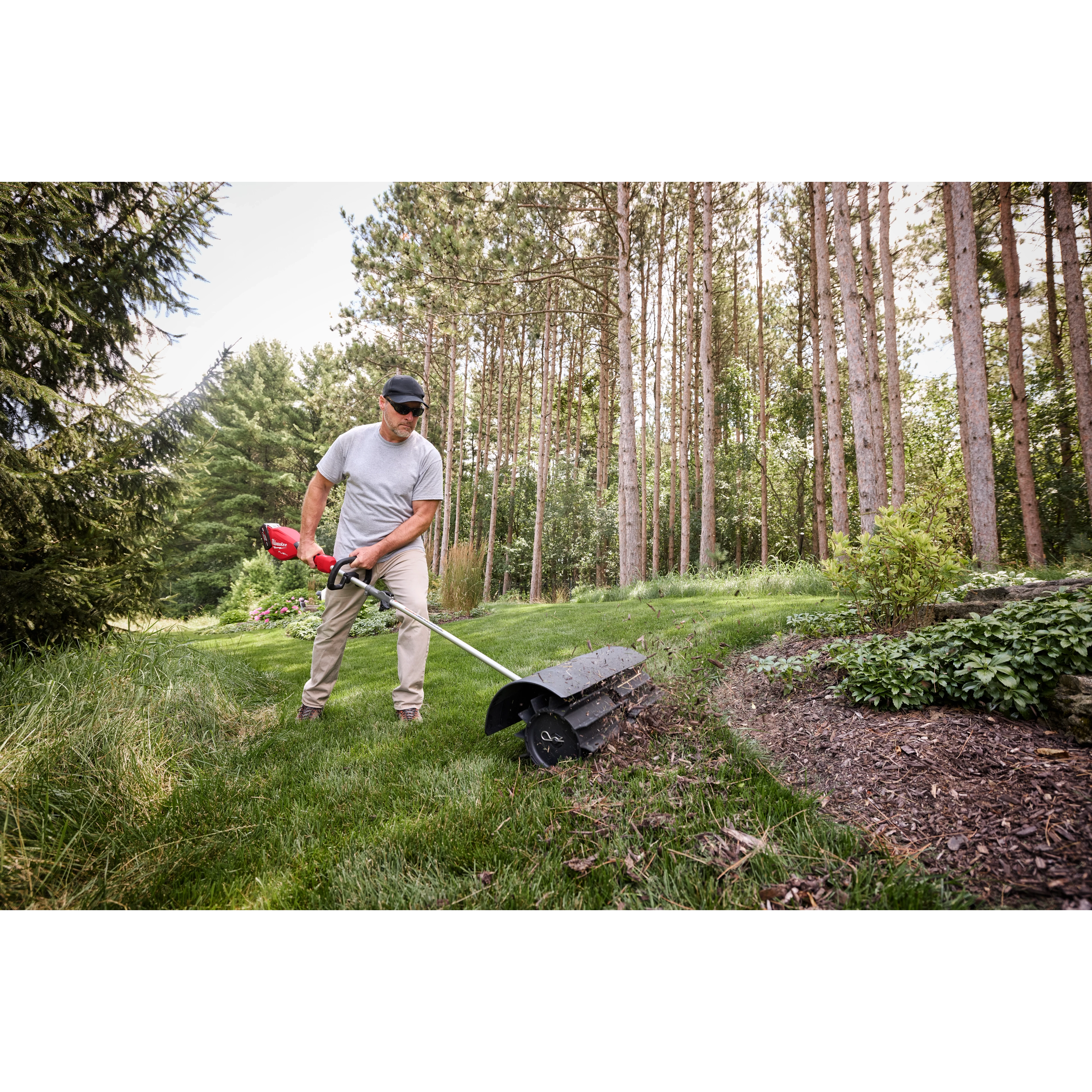 Person using a power tool to maintain a garden, surrounded by lush greenery and tall trees.