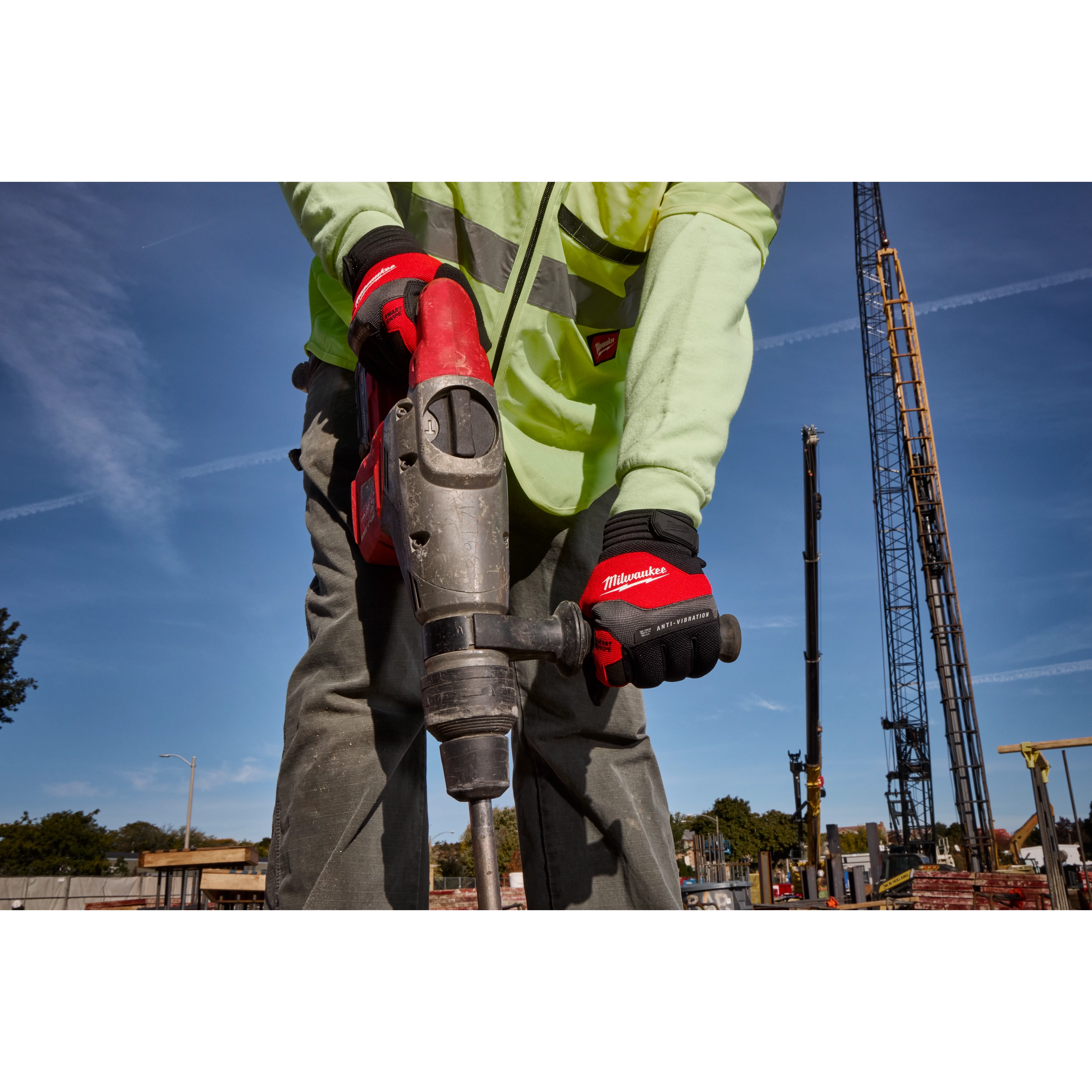 A construction worker wearing Anti-Vibration Work Gloves operates a power tool on a construction site. The worker is dressed in a reflective jacket and grey pants, and the background shows construction cranes and a clear sky.