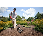 Person using a soil tiller in a garden surrounded by blooming flowers and bright green foliage.