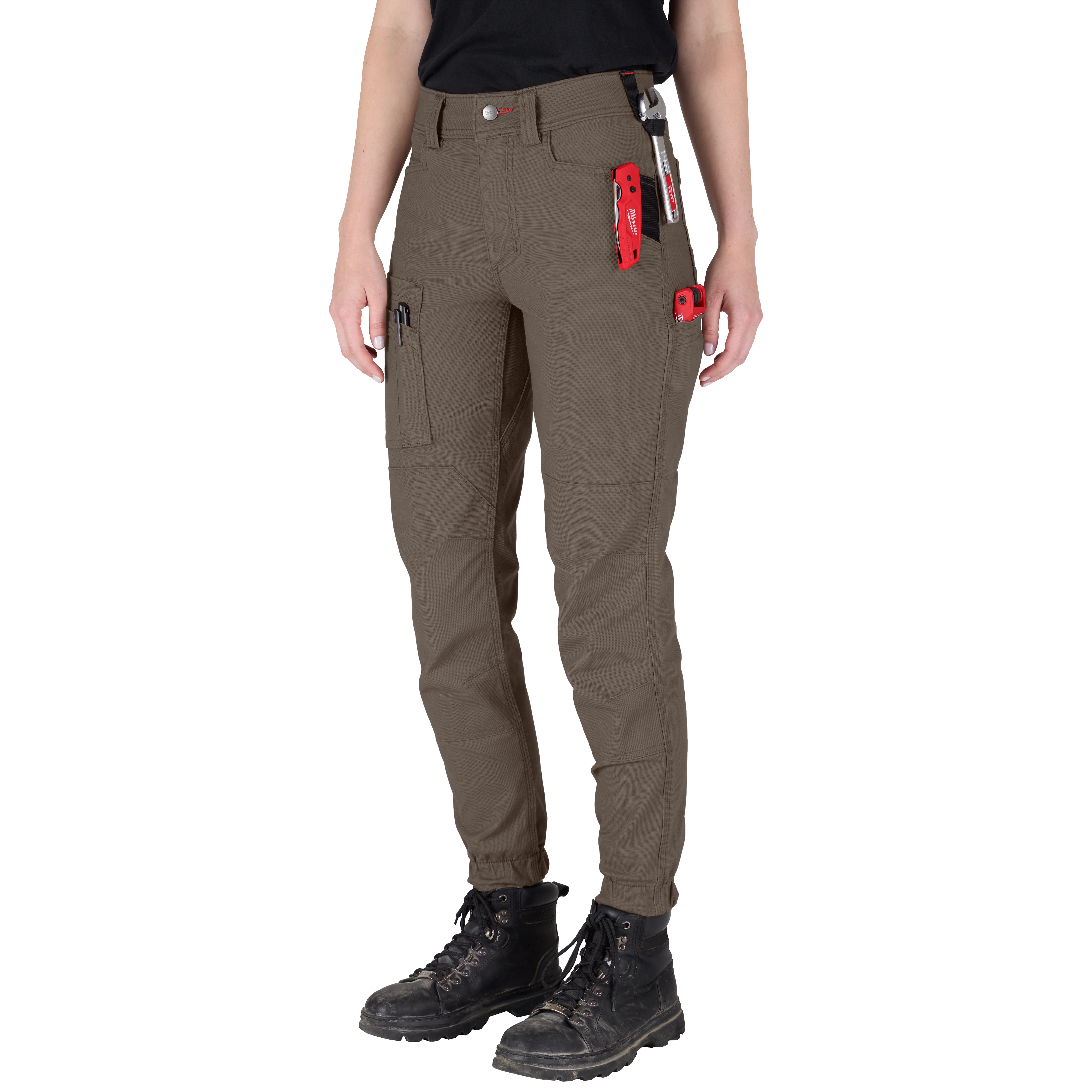 A person is wearing Women's GRIDIRON™ Double Knee Jogger Pants - Brown. The pants have multiple pockets and tools attached, such as a red utility knife, and are paired with black boots.