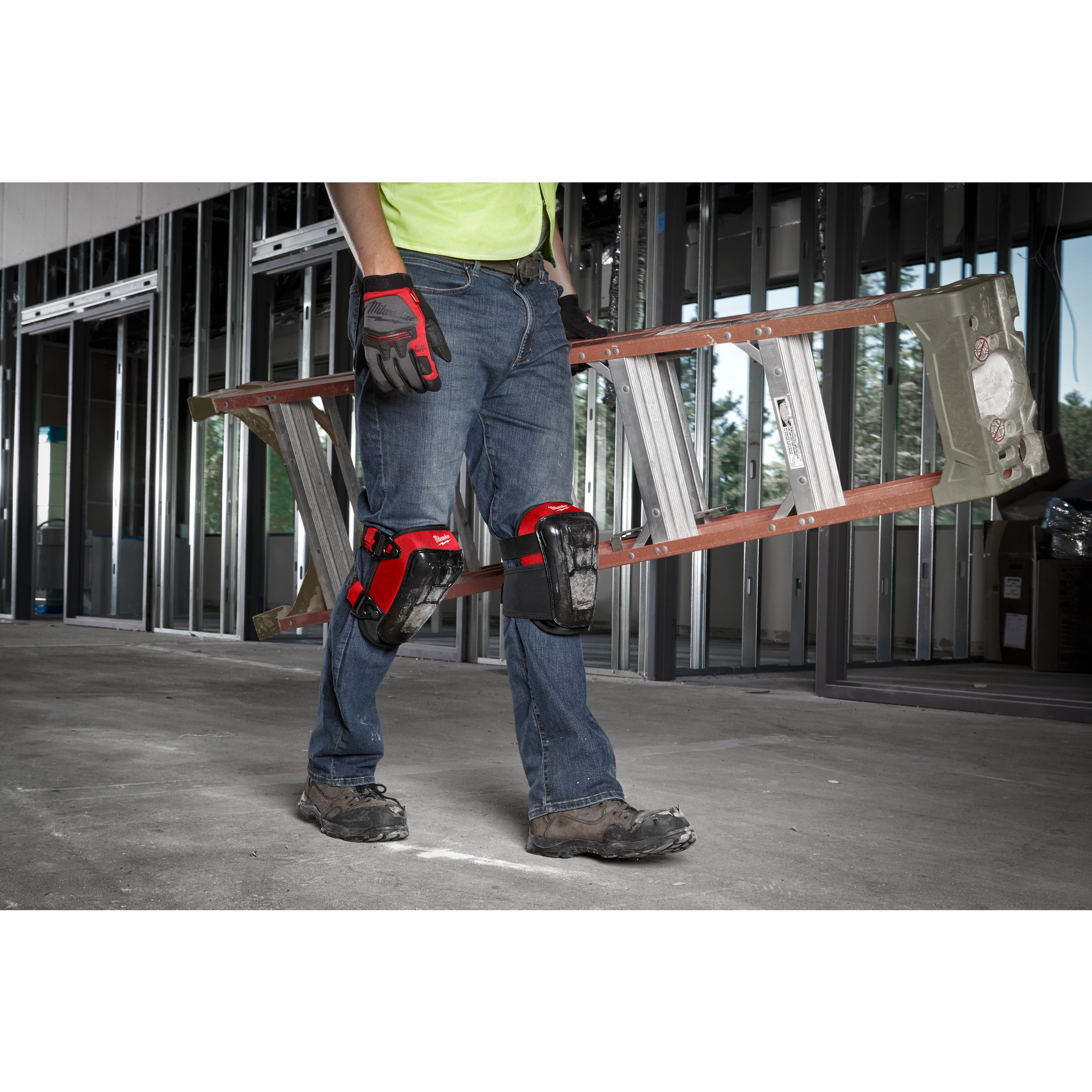 Person wearing knee pads and gloves, carrying a ladder inside a partially constructed building.