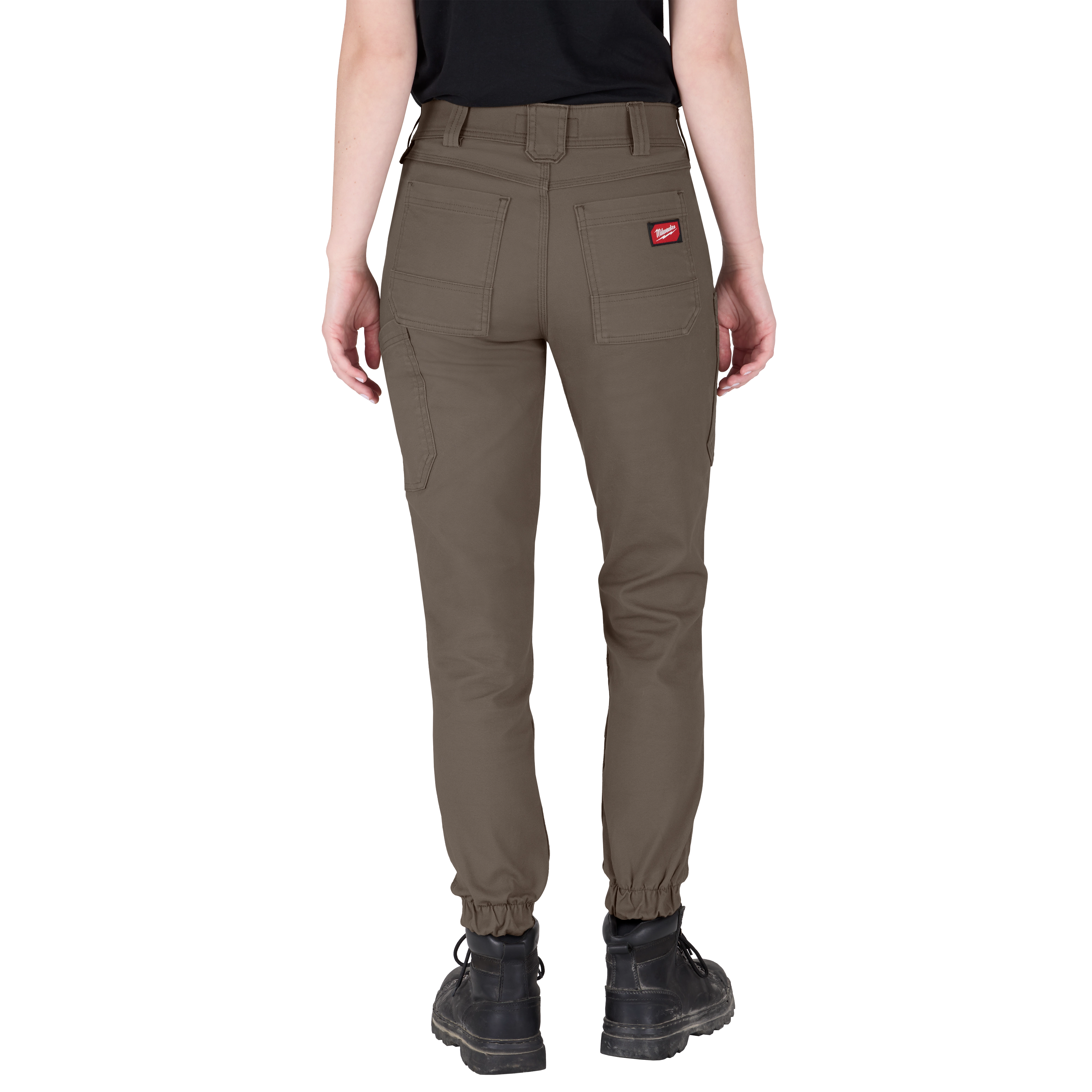 A person wearing Women's GRIDIRON™ Double Knee Jogger Pants - Brown. The brown jogger pants feature multiple pockets, a red logo patch on the back pocket, and elastic cuffs at the ankles. The person is shown from the back, wearing black boots.