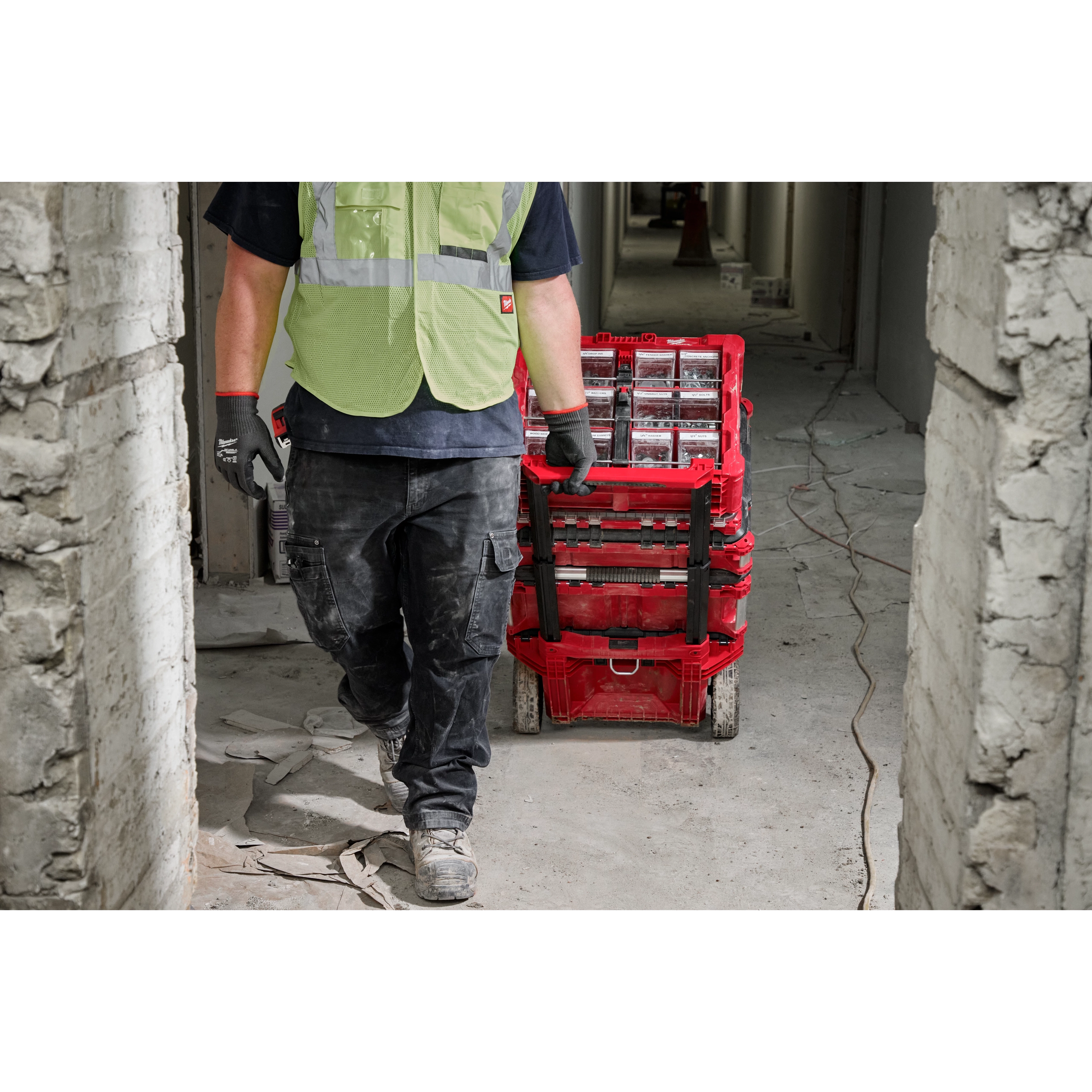 A construction worker in a green safety vest and black gloves is pulling a red cart filled with tools and materials through a narrow passage of a partially constructed building. The floor is covered with debris and electrical wires. The person’s face is not visible.